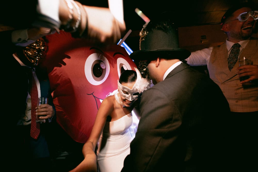 La novia, con antifaz plateado, baila con el novio, que lleva sombrero de copa, junto a un corazón hinchable rojo en la fiesta · Cupido debe morir