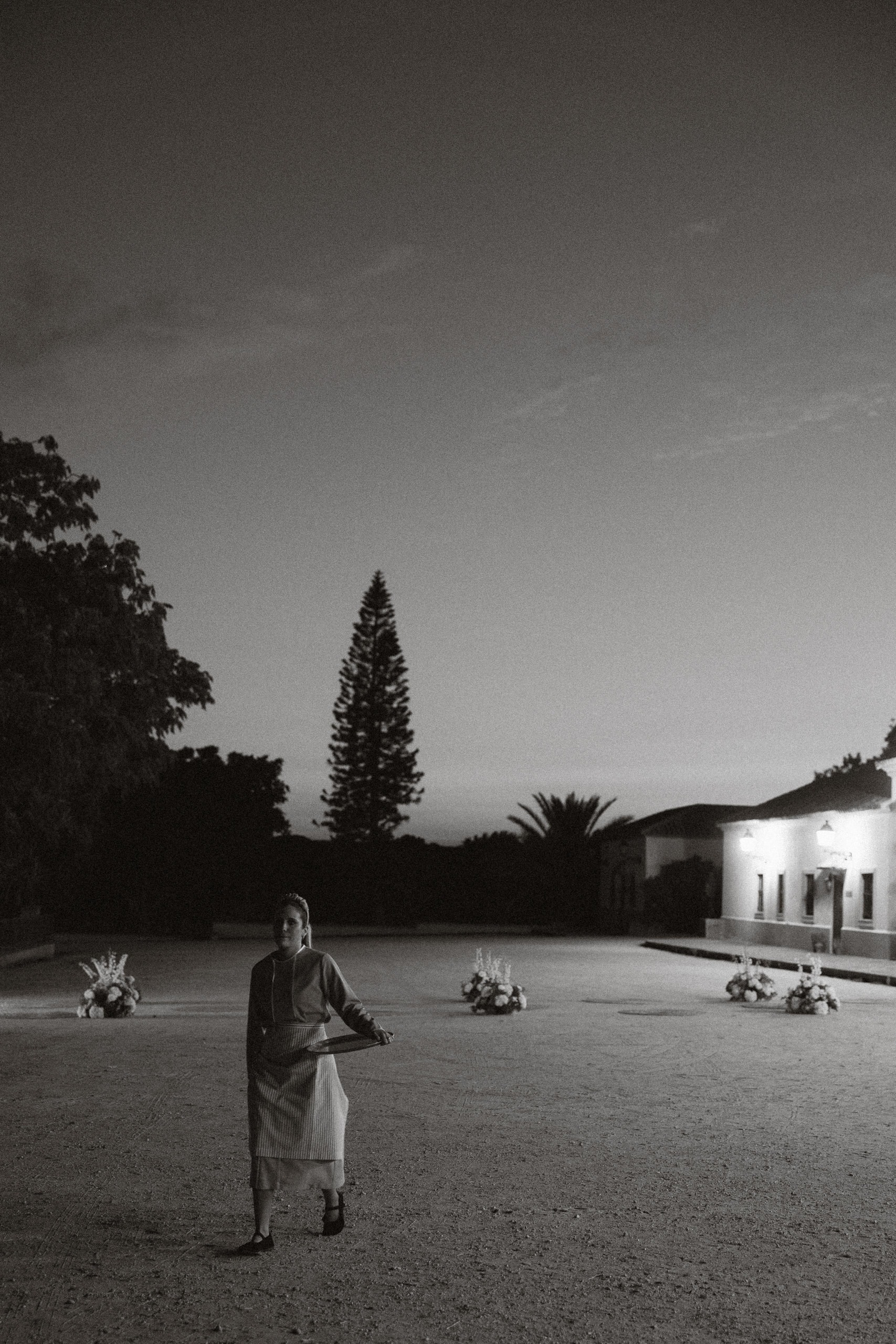 Una camarera con una bandeja cruza una explanada al anochecer, con centros de flores y una casa iluminada al fondo, en blanco y negro · Cupido debe morir