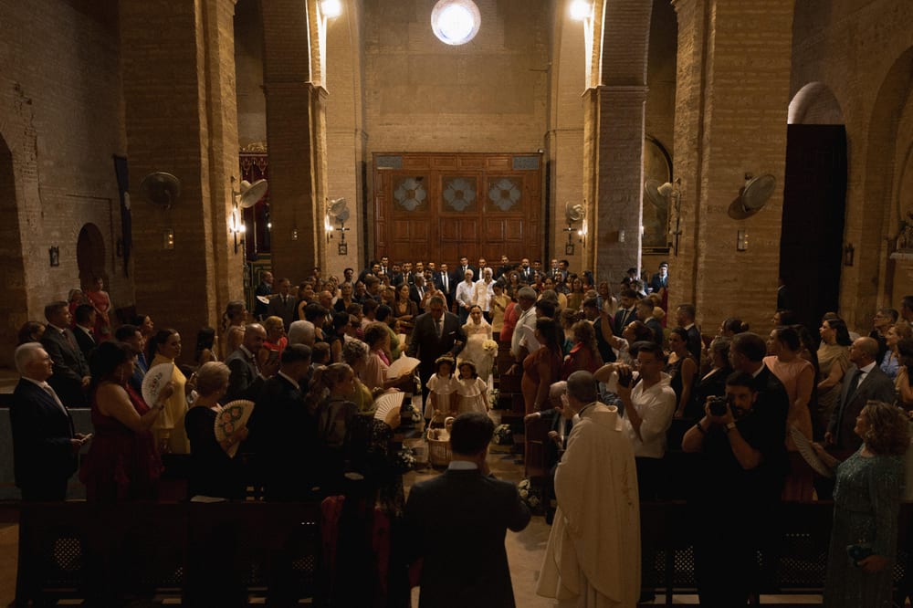 La novia entra del brazo de su padre por la nave de la iglesia, entre invitados con abanicos y dos niñas de arras delante · Cupido debe morir
