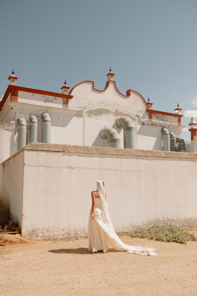 La novia, sola, ante la tapia blanca de la hacienda, con la cola del vestido extendida en el suelo · Cupido debe morir