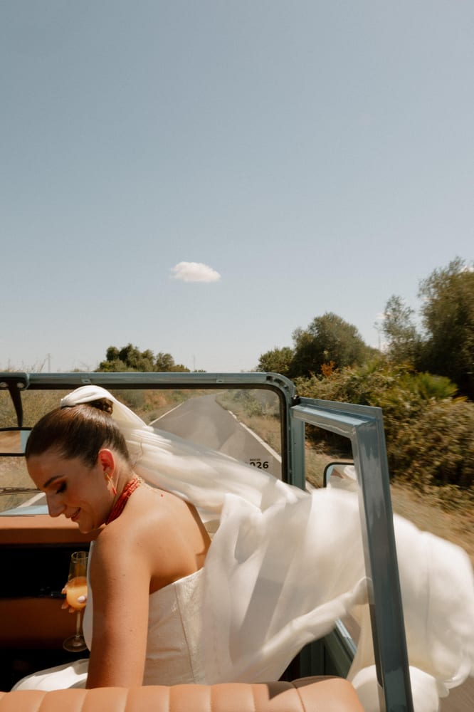 La novia, con velo y ramo, sentada en el Land Rover descapotable entre olivos · Cupido debe morir