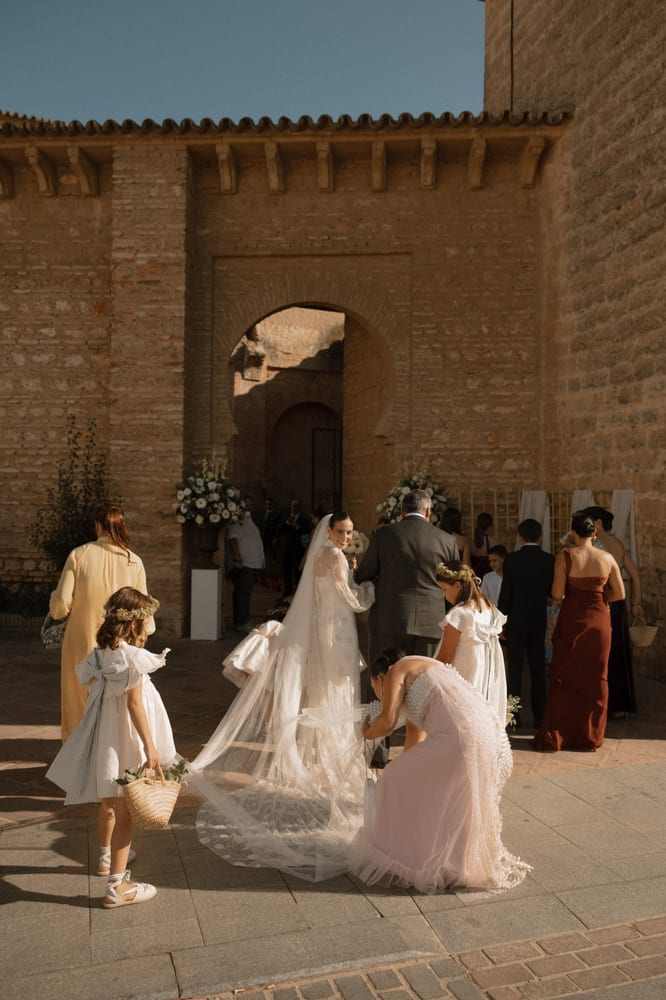 La novia mira sonriendo por encima del hombro, del brazo de su padre, ante el arco de ladrillo de la iglesia mientras una dama de honor le recoge el velo · Cupido debe morir