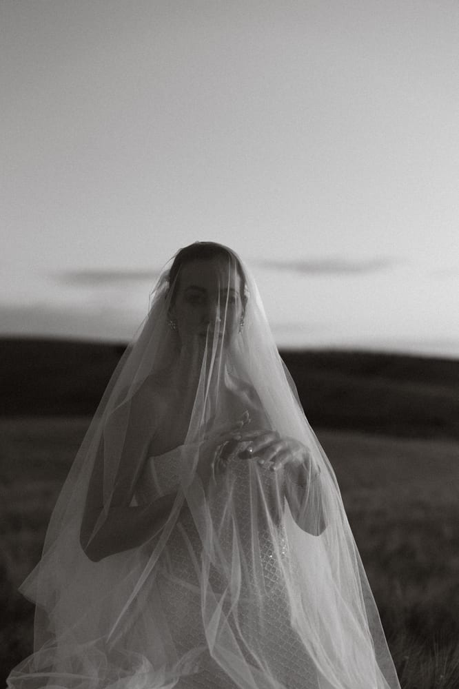 La novia, con el rostro cubierto por un largo velo de tul, sobre un campo al atardecer, en blanco y negro · Cupido debe morir
