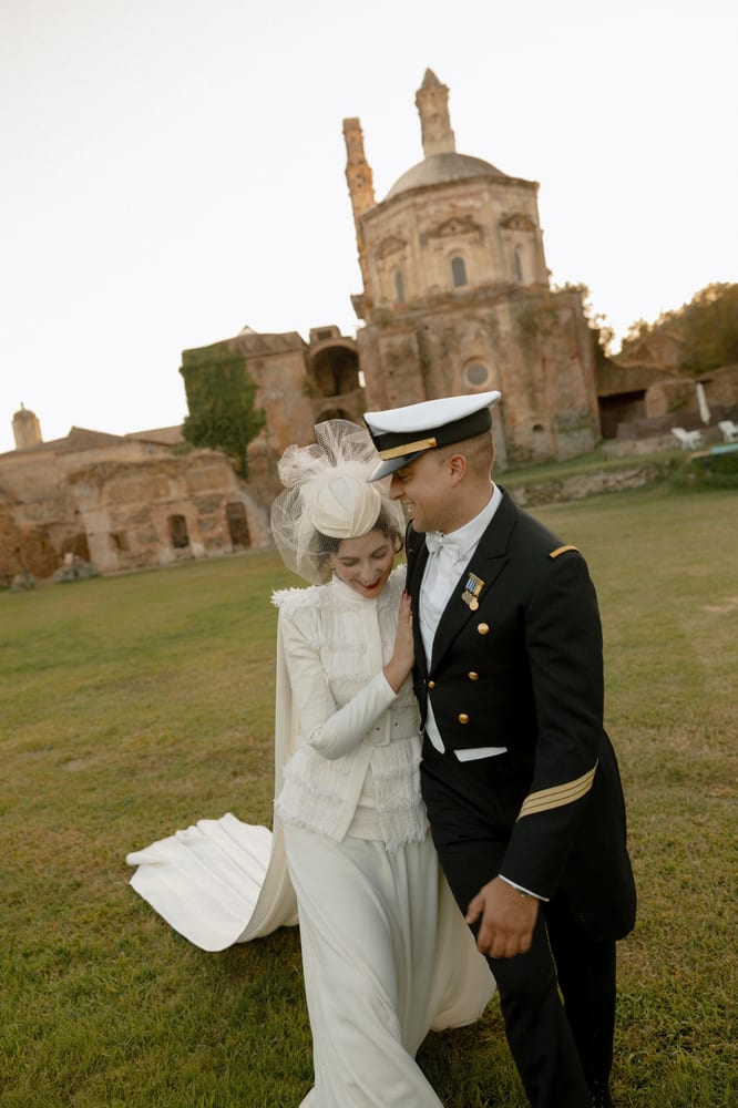 Los novios caminan abrazados sobre la hierba al atardecer, él con uniforme de gala blanco y negro y ella con tocado de redecilla, con un monasterio en ruinas al fondo · Cupido debe morir