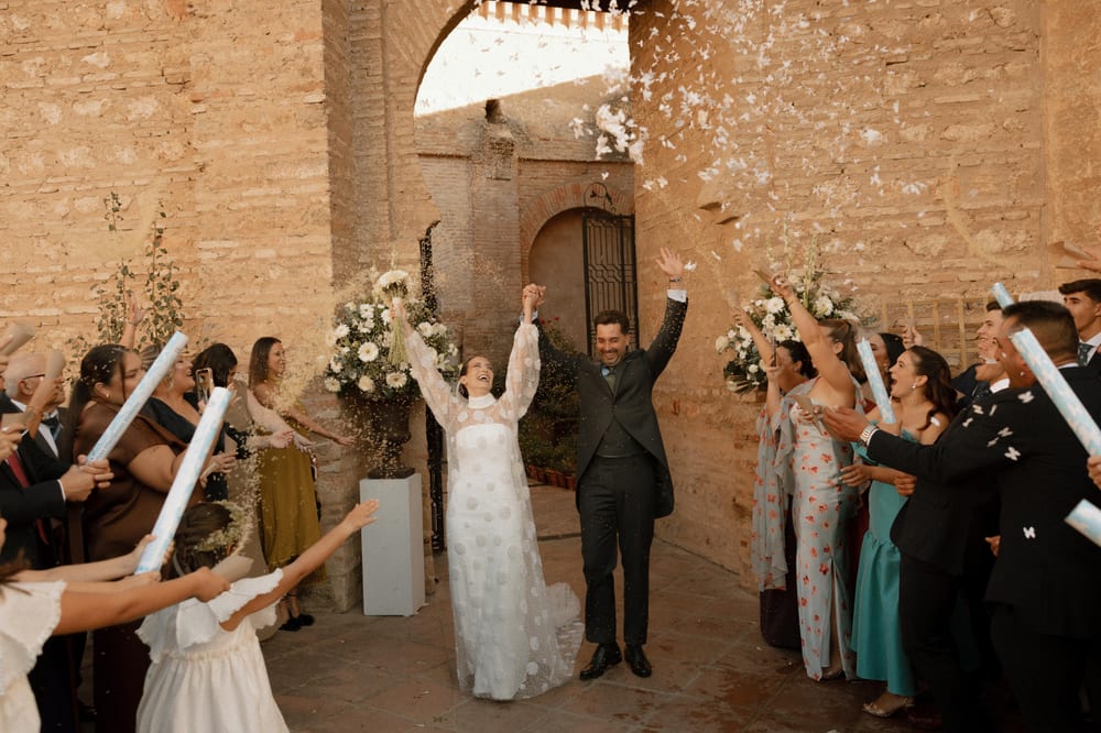 Los novios salen de la iglesia con los brazos en alto entre invitados que lanzan arroz y confeti de mariposas blancas · Cupido debe morir