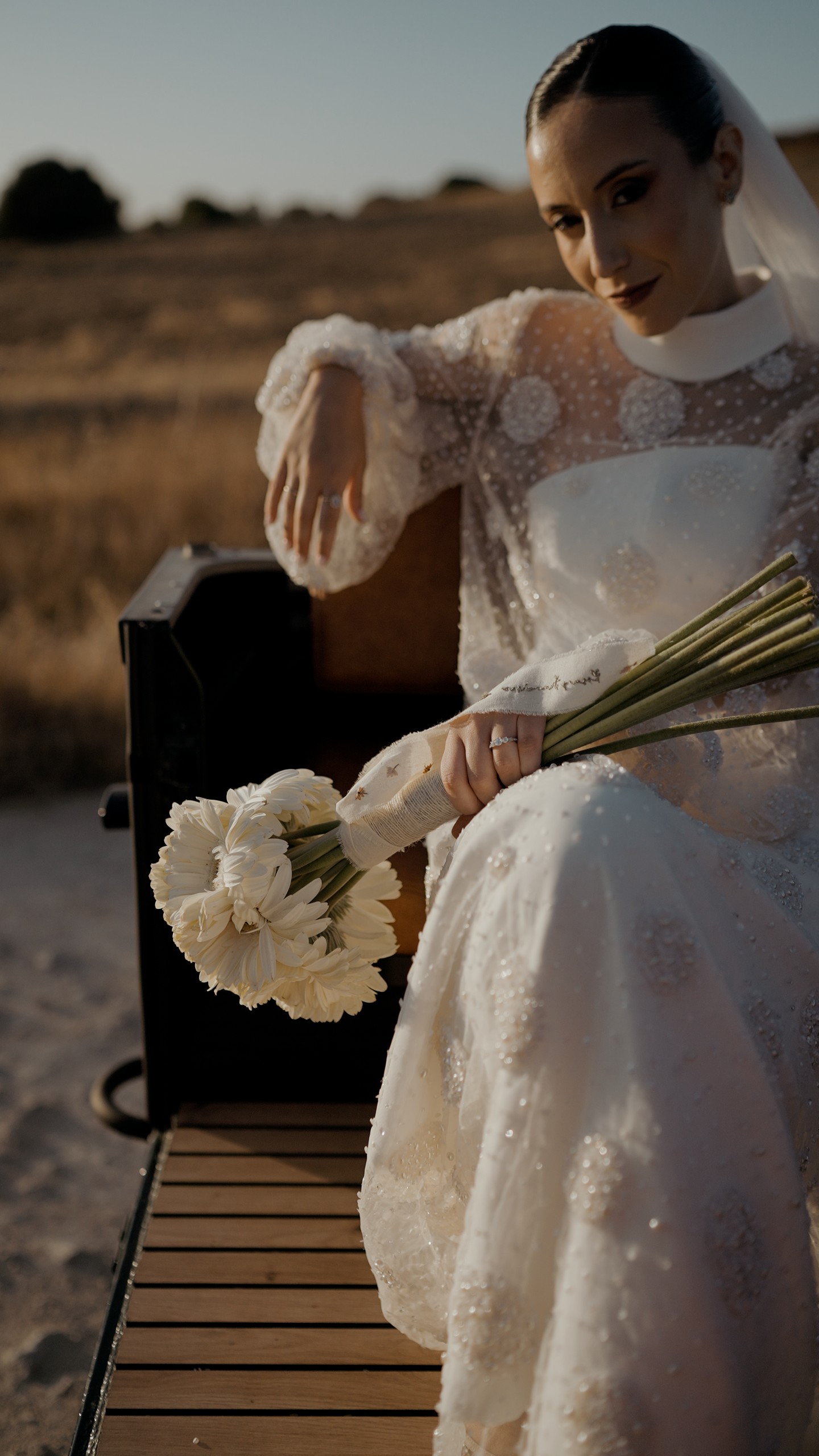 Rosa in her Victoria dress, sitting on the back of a vintage car with her bouquet of white gerberas