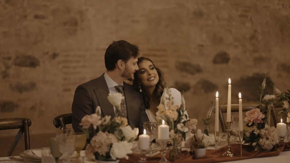 Juan y May, mejilla con mejilla en su mesa, entre velas blancas y flores melocotón