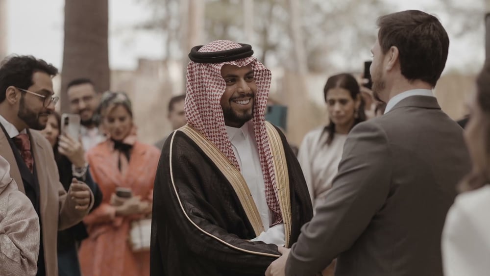 Juan saluda a un invitado con traje tradicional saudí, un bisht negro sobre una thobe blanca y una ghutra de cuadros rojos, entre invitados sonrientes