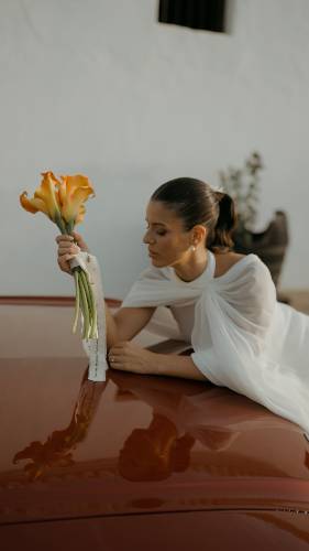 Rocío con su ramo de calas naranjas, apoyada en el capó granate del coche de la boda