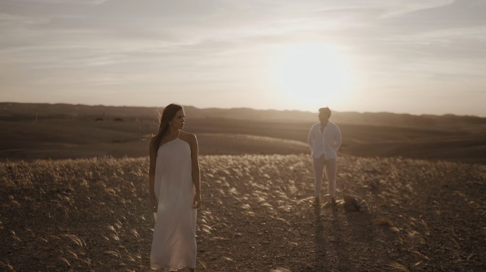 Patri and Juanma, in white, standing apart in a field of golden grass at sunset, during their pre-wedding in Morocco