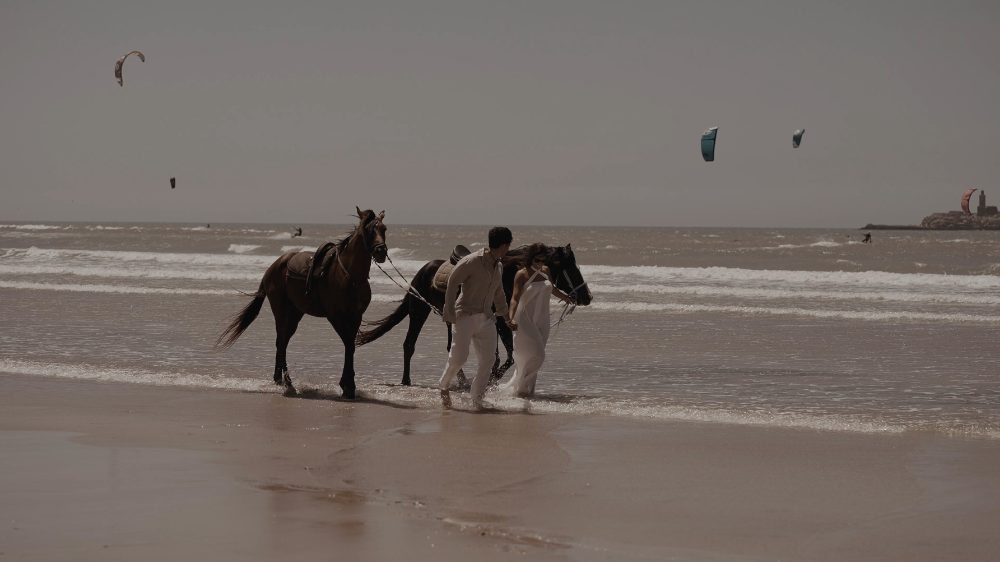 Dos caballos en la orilla de una playa, con dos personas de blanco y cometas de kitesurf al fondo, durante la preboda en Marruecos