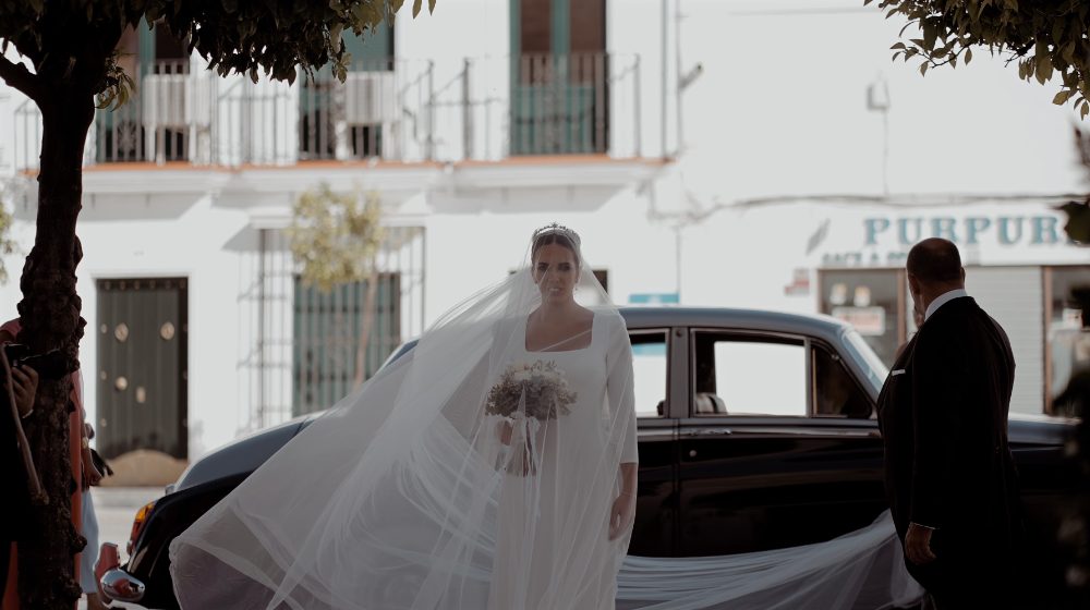 Patri, her long veil in the wind, beside a black classic car on a whitewashed village street