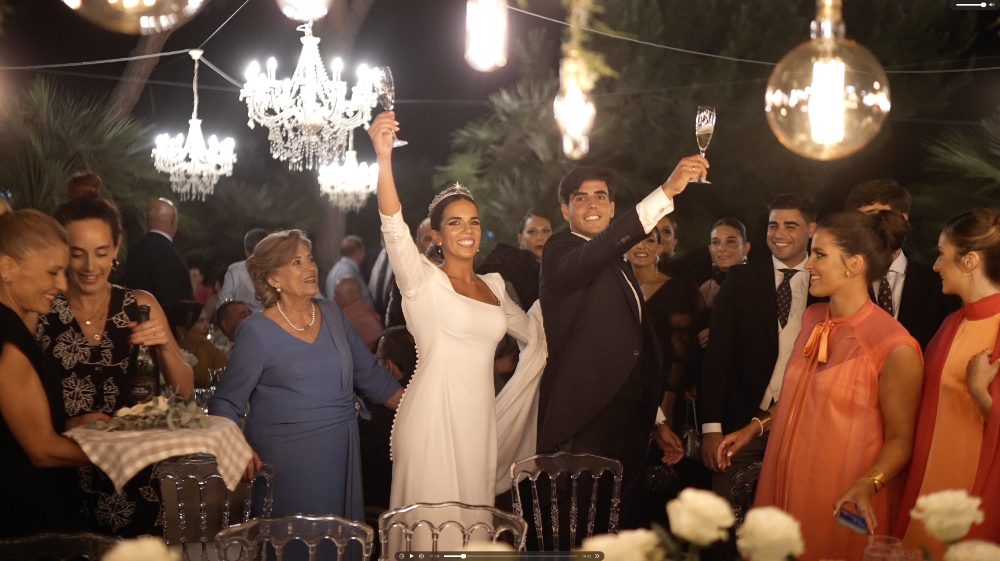 Patri, Juanma and the guests raise their glasses in a toast under hanging lamps, in the garden