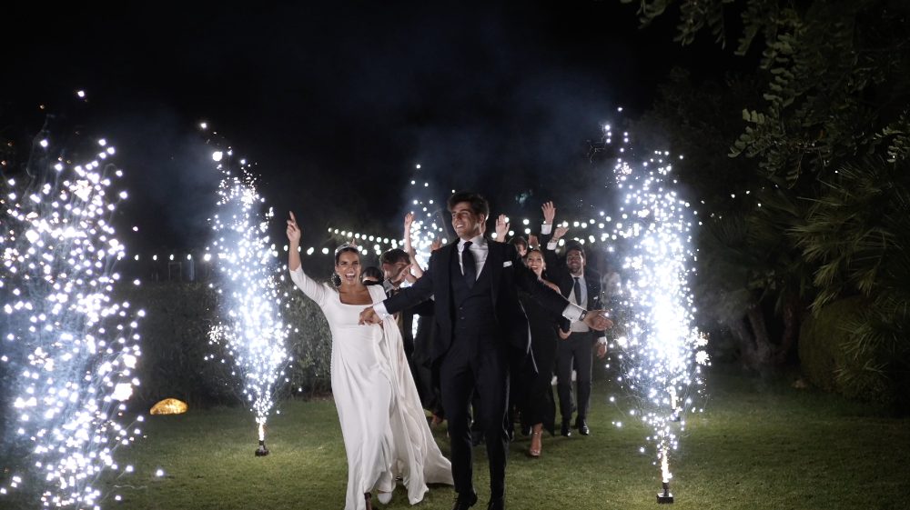 Patri and Juanma walk into the garden at night between sparkler fountains, their guests behind them