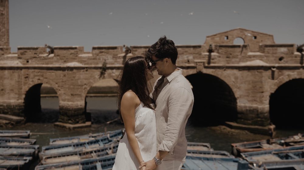 Patri and Juanma, about to kiss, in front of a wall with arches and a harbour full of blue boats, in their pre-wedding in Morocco
