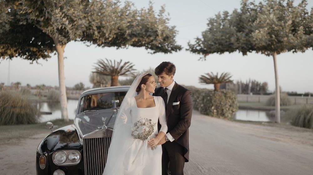 Patri and Juanma smile beside a black classic car, with the lake and palm trees behind them