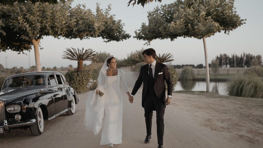 Patri and Juanma walk hand in hand beside the classic car, with the lake and palm trees in the background