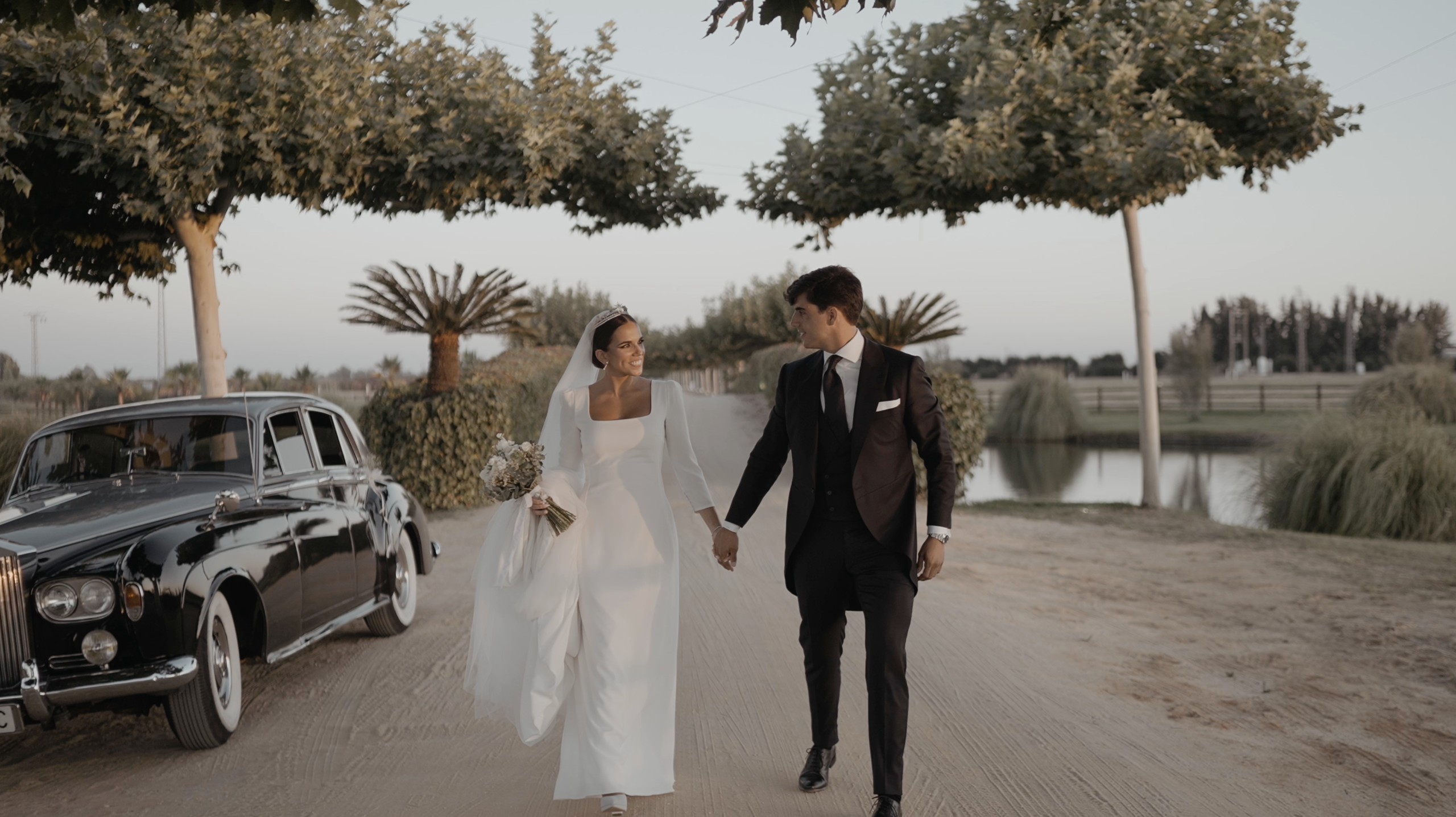 Patri y Juanma, de la mano, caminan junto al coche clásico con el lago y las palmeras al fondo