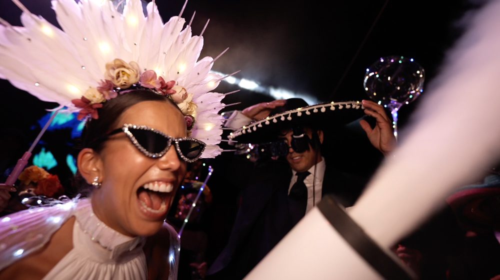 Patri, in a white feather headdress and rhinestone sunglasses, laughs in the middle of the hora loca, with Juanma behind her in a sombrero