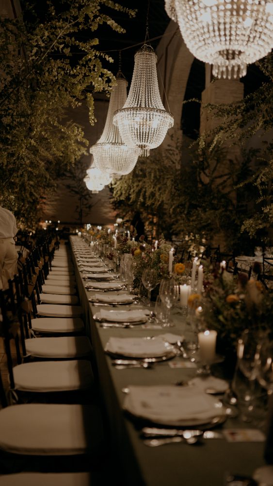 A very long table under chandeliers among greenery, at night · Gearmedia