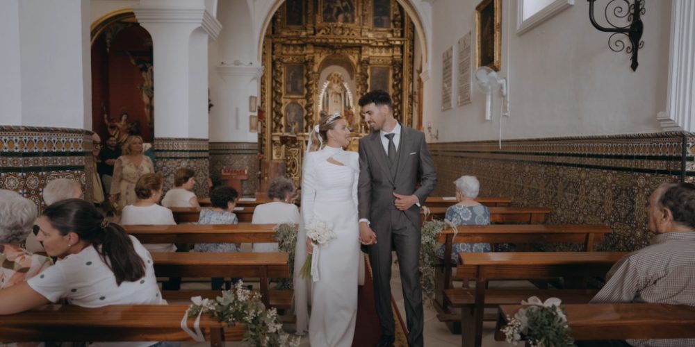 Alicia and Edu holding hands in the nave of the Ermita de Nuestra Señora de Belén, guests on the wooden pews and a gilded altarpiece behind them