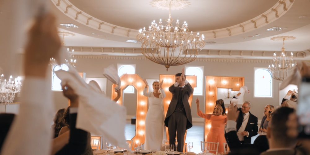 Alicia and Edu entering the dining hall under a crystal chandelier, guests waving napkins around them