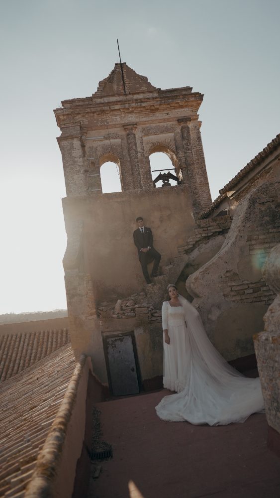 The bride in white, her veil spread out, and the groom sitting on the ruined tower, at a wedding at Convento de la Luz (Huelva) · Gearmedia