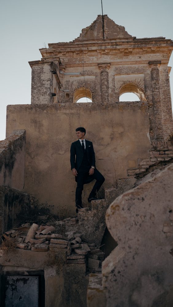 The groom in a dark suit standing on the ruined tower, clear sky behind him · Gearmedia