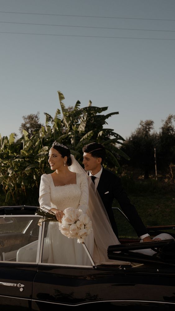 The couple leaning out of a black convertible among banana trees, she with a white bouquet · Gearmedia