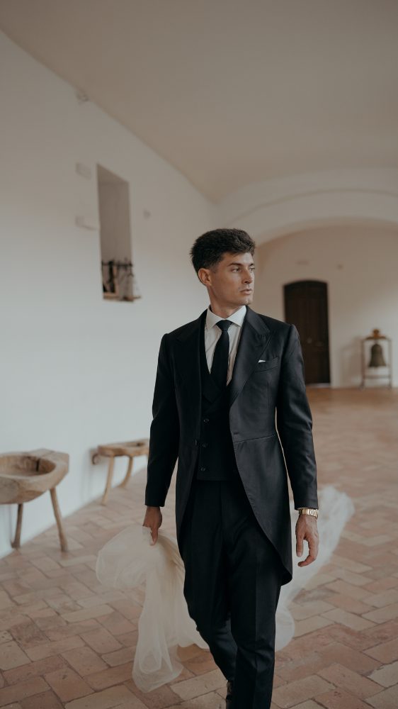 The groom in a morning coat crosses a whitewashed room carrying the dress train in his hand · Gearmedia