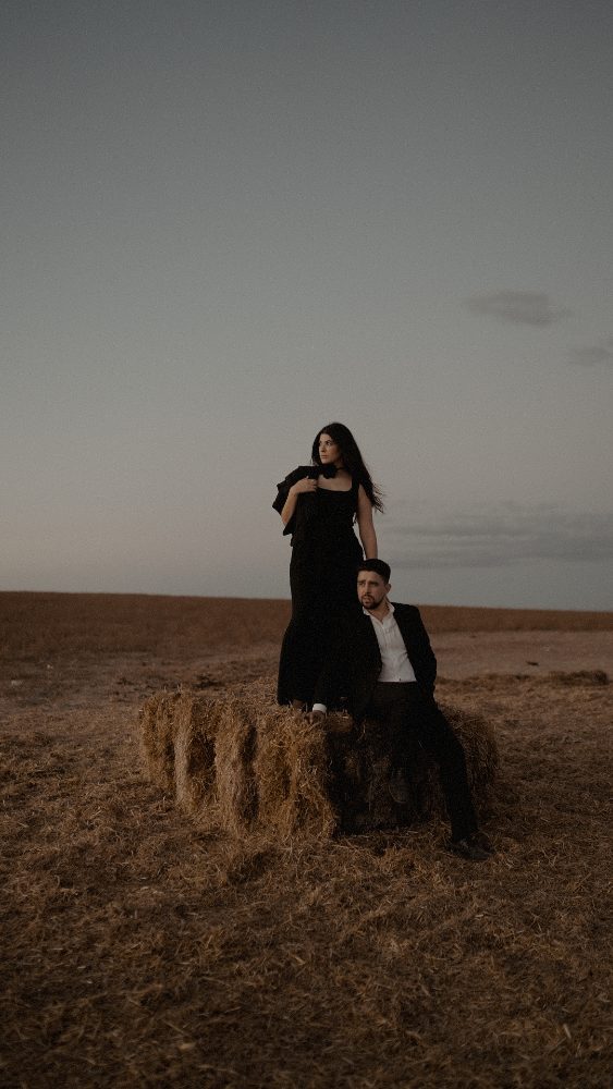 She stands in black and he sits on a hay bale, sunset sky · Gearmedia