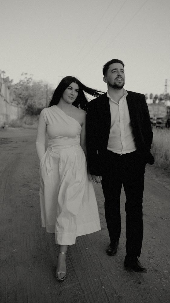 Black and white: the couple walking arm in arm along a dirt path, her hair in the wind · Gearmedia
