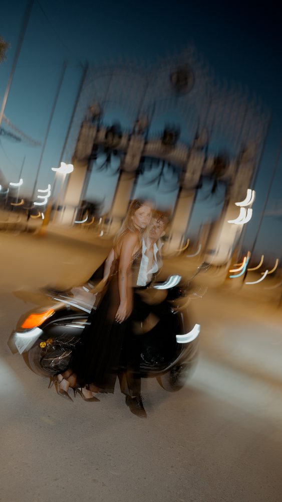 Rocío y Pablo, junto a una moto, de noche, con la portada iluminada de una feria al fondo y las luces en estela