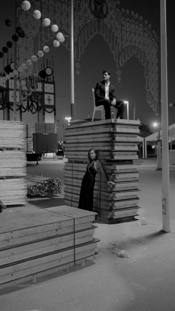 En blanco y negro, Pablo sentado en una silla sobre un montón de tablones y Rocío de pie, apoyada en ellos, bajo la portada iluminada de una feria