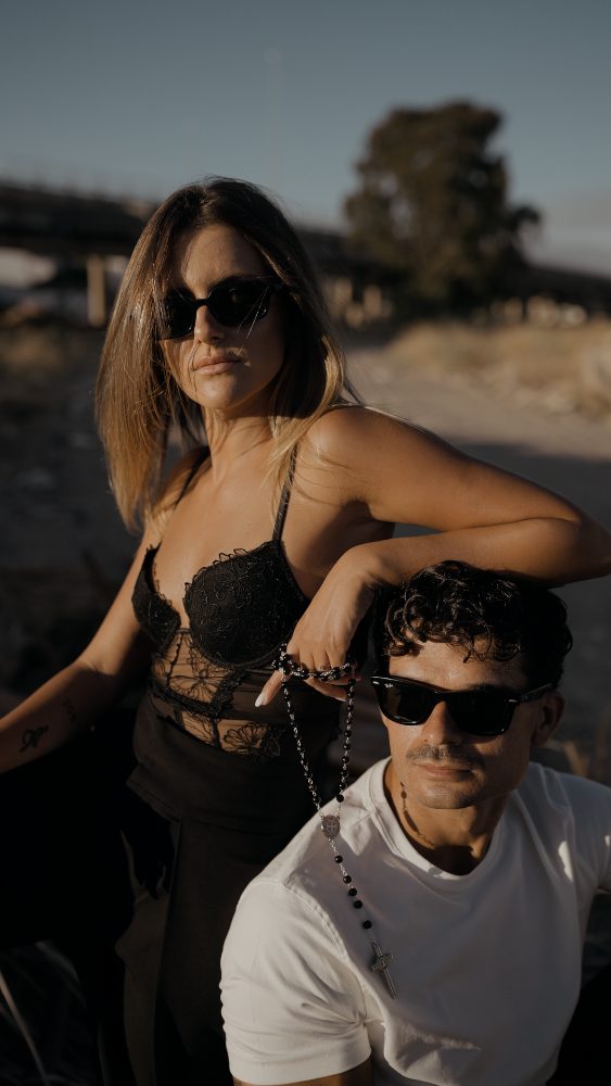 Una mujer con gafas de sol y un hombre con gafas y un rosario al cuello, de camiseta blanca, posan al atardecer en un descampado