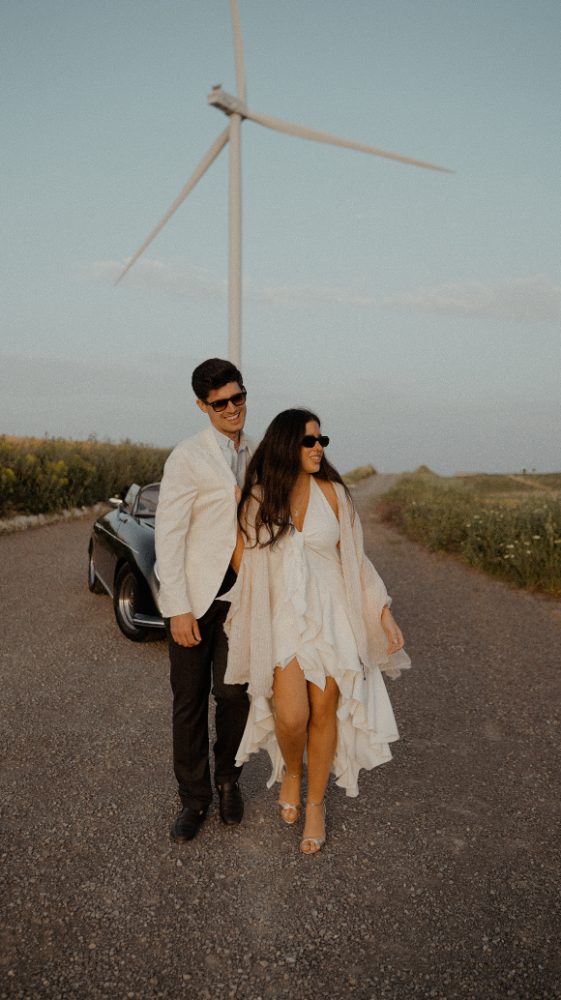 A couple in sunglasses, he in white, she in a short dress, beside a classic car and a wind turbine at sunset · Gearmedia
