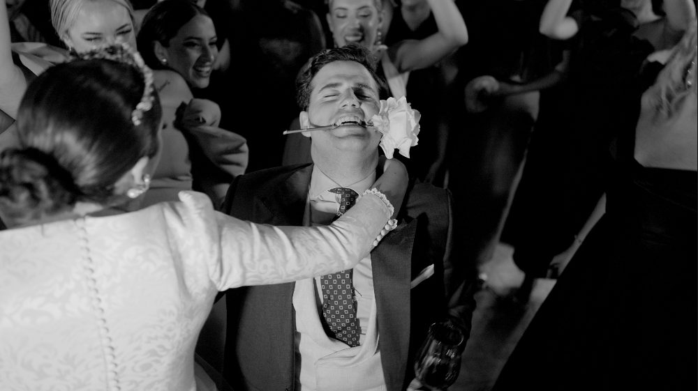 Black and white, mid-party: the bride pulls the groom by his tie among the guests · Gearmedia