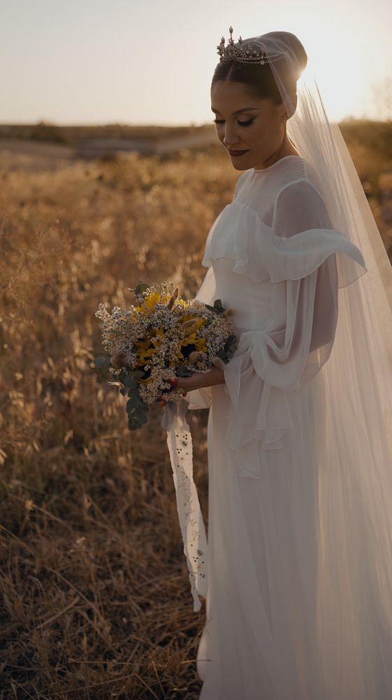 The bride with a crown and veil in a wheat field at sunset, holding wildflowers · Gearmedia