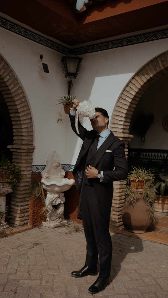 Pablo, de chaqué, se tapa la cara con el ramo blanco de Rocío en alto, en un patio de arcos de ladrillo y paredes blancas