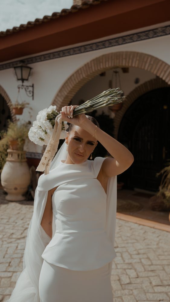 Rocío levanta el ramo de flores blancas sobre la cabeza, con la capa y el velo al viento, en el patio empedrado