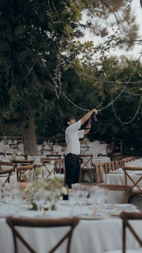 A person raising a camera between the banquet tables, under strings of lights hung from the trees