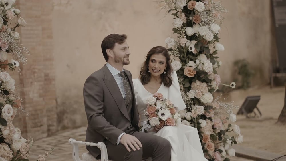 Juan and May sit side by side in front of a tall arch of cream and peach roses, laughing
