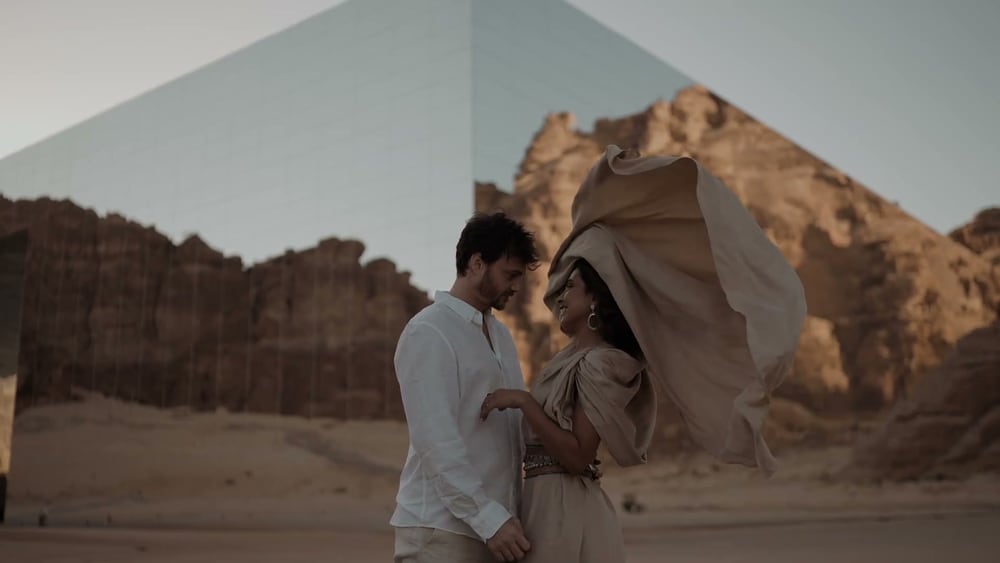 Juan and May in the desert at AlUla, Saudi Arabia, her cape lifted by the wind, a mirrored building and sandstone cliffs behind them