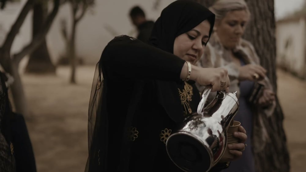 A woman in black pours Arabic coffee from a silver pot under the trees, a guest beside her