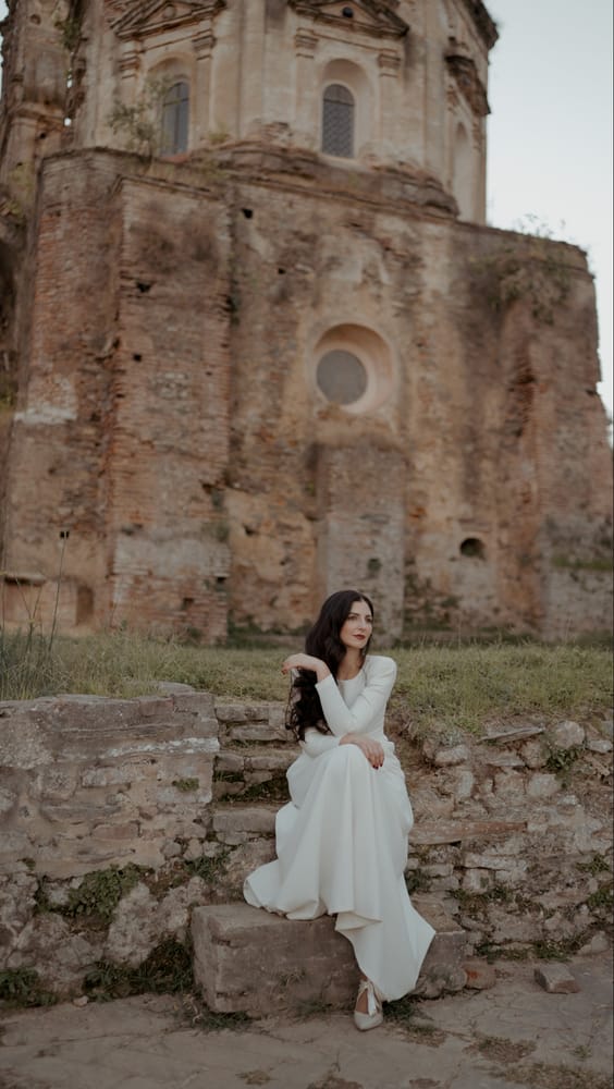 Novia sentada en un muro de piedra ante una iglesia en ruinas, en La Cartuja de Cazalla, Sevilla