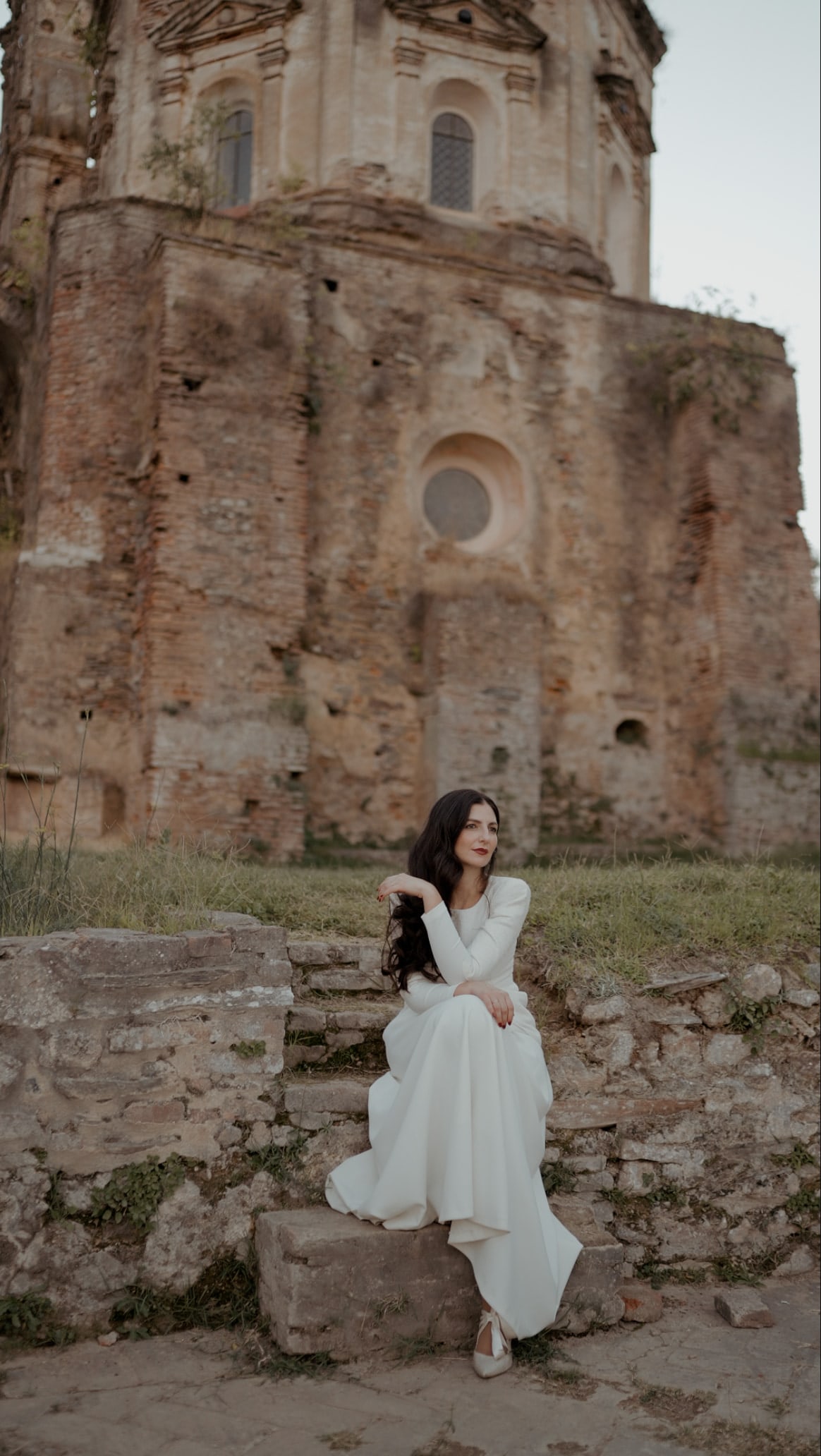 Novia sentada en un muro de piedra ante una iglesia en ruinas, en La Cartuja de Cazalla, Sevilla