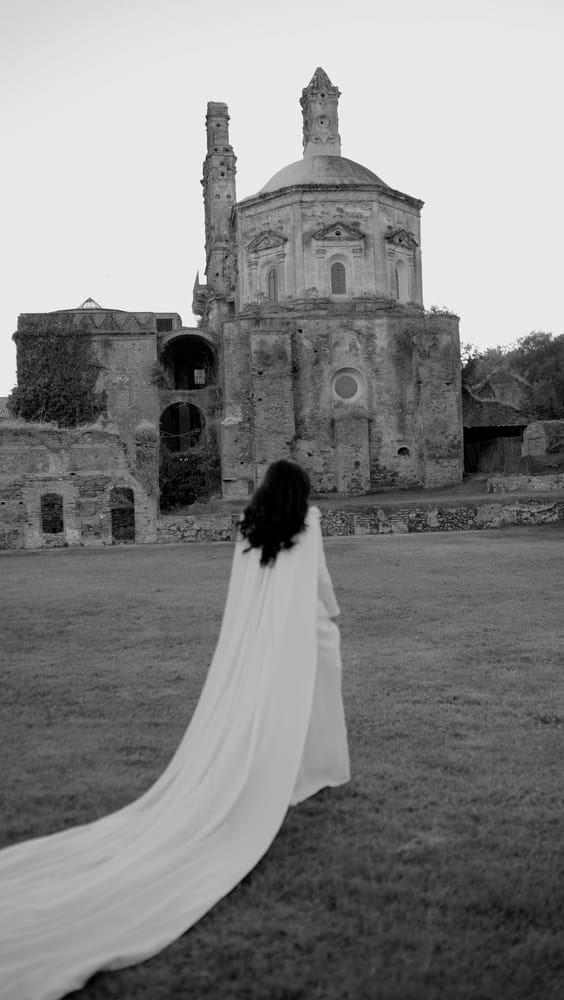 Novia con una capa larga caminando hacia una iglesia en ruinas, en La Cartuja de Cazalla, Sevilla